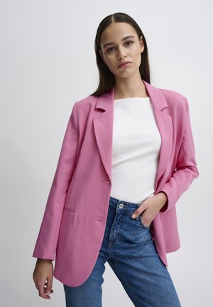 Roze blejzer od glatkog materijala, s rebrastim ovratnikom, dva prednja džepa i ležernim krojem, uparen s plavim trapericama.