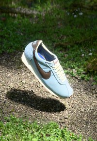 Zapatilla azul claro con parte superior texturizada, logotipo de swoosh marrón, suela beige y cordones crema, situada sobre un fondo verde natural.