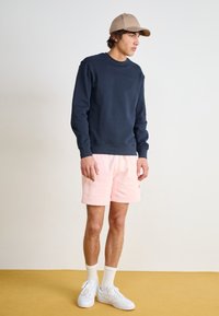 Marineblå bomulds-sweatshirt, lyserøde bomulds-shorts. Hvide sneakers, strømper i ankellængde. Stående på et gult gulv, minimalistisk baggrund, afslappet pasform.