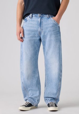 Persona che indossa jeans larghi azzurri chiaro con le mani nelle tasche e sneakers nere, in piedi davanti a uno sfondo semplice.