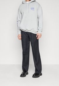 Felpa grigia in cotone con tasca a marsupio anteriore e logo blu, abbinata a pantaloni scuri e scarpe nere. Design semplice con una vestibilità rilassata.
