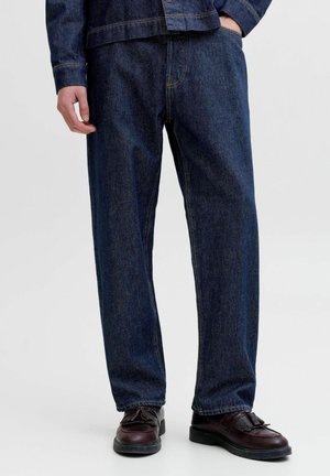 Homme portant un jean en denim bleu foncé, une veste en denim foncé et des chaussures en cuir marron foncé, debout sur fond blanc.