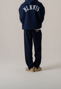 Navy blauwe hoodie met witte "BLKVIS" letters op de achterkant, gecombineerd met bijpassende broek. Zacht materiaal en een relaxte pasvorm, met een zijzak.