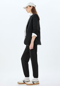Zwarte blazer met een wit shirt eronder, zwarte taps toelopende broek en witte sneakers met zwarte accenten en een bruine zool. Lichtgrijze pet.