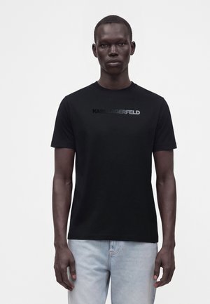 Uomo che indossa una maglietta nera a maniche corte con il testo "KARL LAGERFELD" e jeans azzurri, in piedi davanti a uno sfondo bianco uniforme.