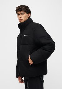 Junger Mann trägt eine schwarze Steppjacke mit hohem Kragen, eine Hand in der Tasche, leicht nach links gewandt vor einem schlichten hellen Hintergrund.