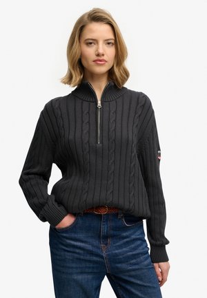 Kobieta w czarnym swetrze warkoczowym z półzamkiem przy kołnierzu, niebieskich dżinsach i brązowym plecionym pasku, stoi z jedną ręką w kieszeni.