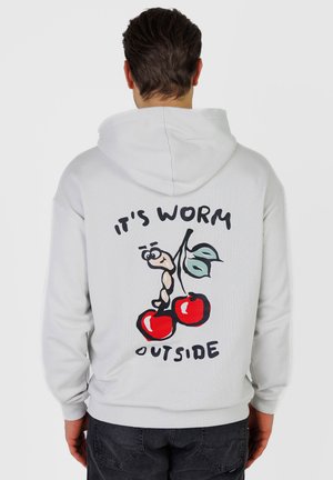 Mand iført hvid hættetrøje med tegneserieorm, der klatrer op ad to røde kirsebær og tekst "It's worm outside" på ryggen.