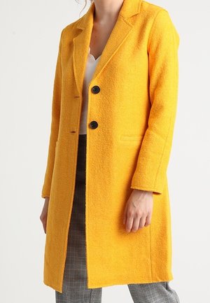 Kobieta ubrana w jasny żółty teksturowany wełniany płaszcz z czarnymi guzikami, biały top i szare spodnie w kratę na jednolitym tle.