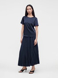Femme aux longs cheveux foncés portant un haut bleu marine à manches courtes, une jupe longue plissée et des sandales noires à talons, debout devant un fond uni.