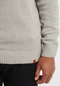 Grauer Strickpullover mit gerippten Bündchen und Saum, ausgestattet mit einem kleinen braunen Lederlogo-Patch nahe dem Saum. Weiche Textur und figurbetontes Design.
