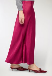 Gonna lunga in satin fucsia con silhouette fluida, caratterizzata da una spacco laterale. Abbinata a mule rosse con punta a punta e fiocco decorativo.