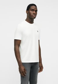 Hombre de pie vistiendo una camiseta blanca lisa de manga corta y jeans grises contra un fondo claro y liso.