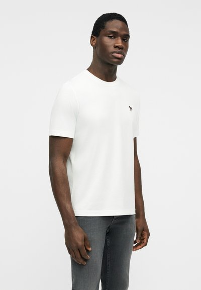 Άνδρας που στέκεται φορώντας απλό λευκό κοντομάνικο μπλουζάκι και γκρι τζιν, σε απλό ανοιχτόφωτο φόντο.