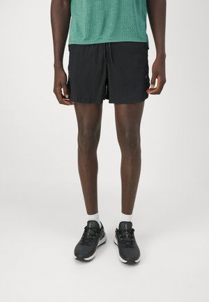 Mann trägt grünes, strukturiertes T-Shirt, schwarze Sportshorts, weiße Knöchelsocken und schwarze Laufschuhe und steht vor weißem Hintergrund.
