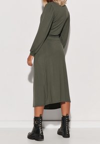 Robe vert olive à manches longues en tissu doux, avec une taille cintrée et une jupe fluide, associée à des bottines noires cloutées.