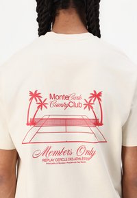 T-shirt color crema con un design grafico rosso, che include palme e un campo da tennis, con la scritta "Monte Carlo Country Club" e "Solo per membri".