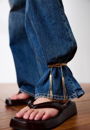 Jeans cuffed at the ankle with a gold chain accessory, worn with black platform flip-flops on a wooden floor.