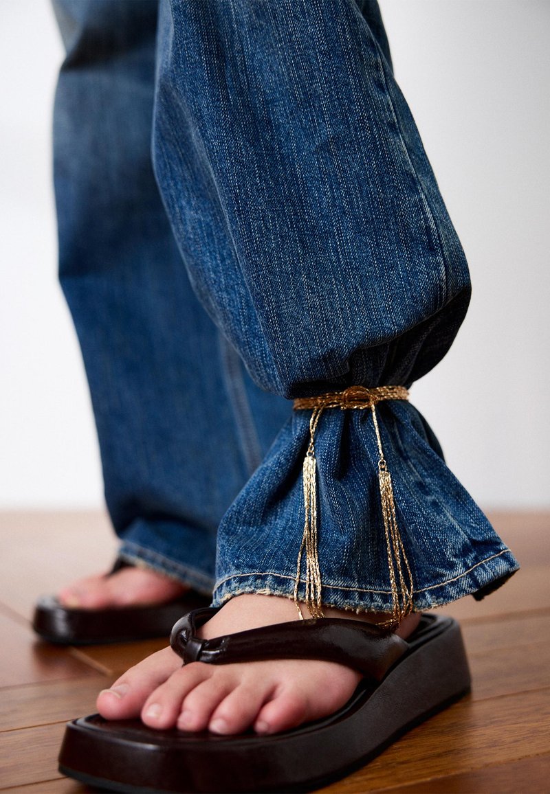 Jeans med opbukket kant ved anklen og en guldkæde som tilbehør, båret med sorte platform-flipflops på et trægulv.
