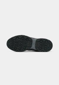 Black athletic shoe sole with rugged tread pattern, featuring grooves and an embossed logo. Material appears rubberized and textured.