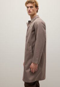 Cappotto trench marrone chiaro con taglio dritto, dotato di colletto, tasche laterali e un tessuto testurizzato. Il capo ha maniche lunghe e un design minimale.