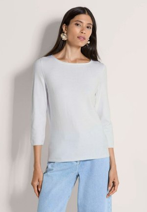 Vrouw draagt een wit topje met driekwartmouwen en lichtblauwe spijkerbroek, gecombineerd met grote pareloorbellen, staand tegen een effen achtergrond.