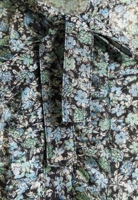 Tejido floral negro con flores azules y verdes que presenta detalles con volantes y un lazo. La textura parece ligera con un brillo sutil.