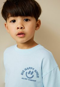 Svetlomodré bavlnené tričko s okrúhlym výstrihom, krátkymi rukávmi a potlačou grafikou s textom "VEĽKÝ ŠŤASTNÝ ÚSMEV" a detailom usmievajúcej sa tváre.