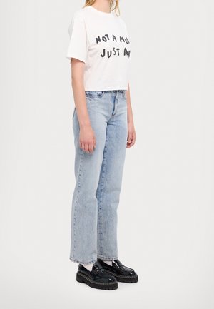 Femme portant un T-shirt court blanc avec du texte noir, un jean droit bleu clair, des chaussettes blanches et des mocassins noirs épais, se tenant de profil.