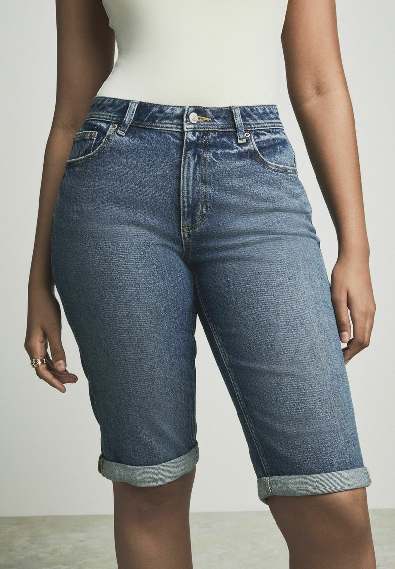 Siniset denim-bermudashortsit, joissa on klassinen viiden taskun muotoilu, käännetyt lahkeet ja nappisuljenta vyötäröllä.