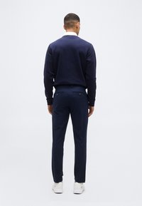 Homme de dos portant un pull marine, un pantalon marine, une chemise blanche à col et des baskets blanches, sur un fond clair uni.