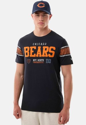 Jonge man draagt een marineblauw Chicago Bears T-shirt met oranje letters en een bijpassende marineblauwe pet met een oranje "C" logo.