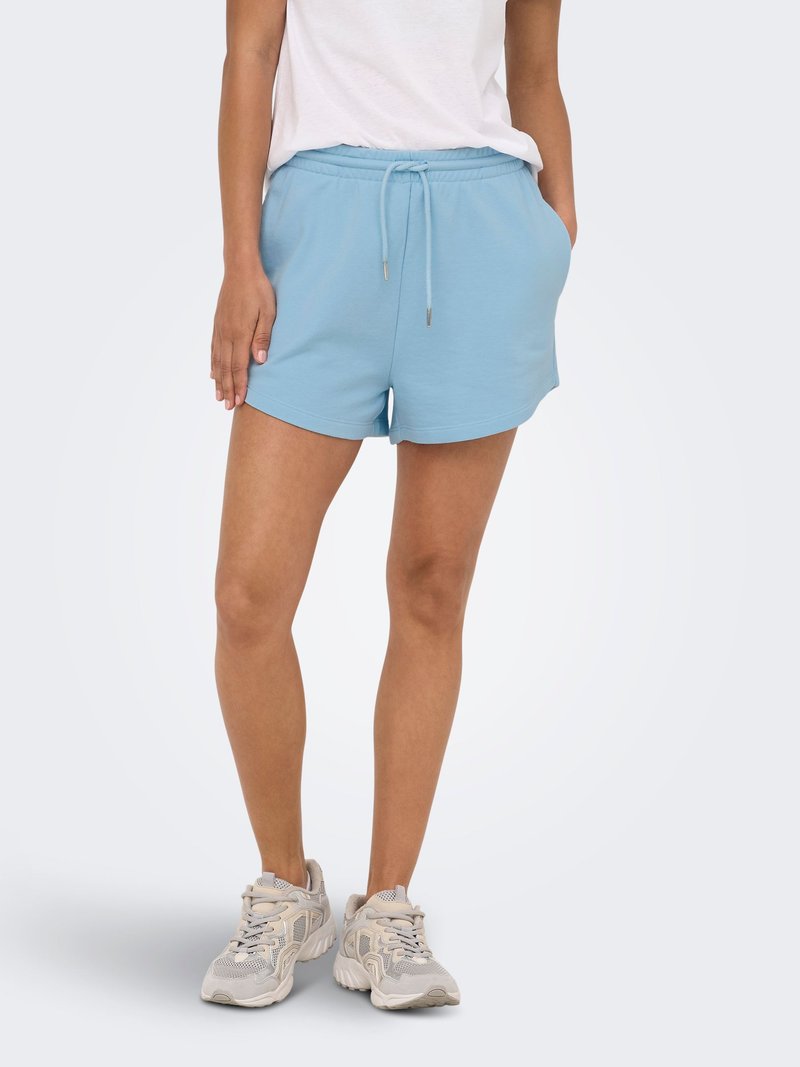 Personne portant un short bleu clair à cordon, un t-shirt blanc et des baskets beiges, debout devant un fond clair uni.