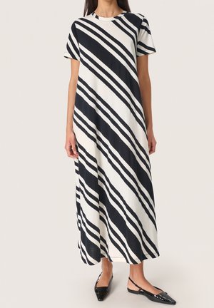Kvinde iført en kortærmet, lang sort og hvid stribet kjole med sorte spidse slingback-flats mod en ensfarvet baggrund.