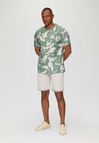 Grünes kurzärmeliges Hemd mit weißem Blättermuster, kombiniert mit hellbeigen Shorts und beigen Sneakers. Weicher Stoff, entspannte Passform.