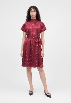 Une femme porte une robe bordeaux à pois blancs, avec un col montant et un lien à la taille, assortie à des chaussures plates noires à bout pointu.