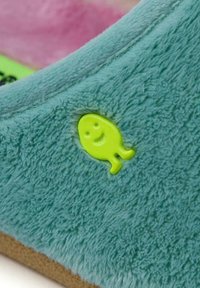 Chausson en peluche turquoise doté d'une texture douce et duveteuse, avec un accent en forme de poisson souriant de couleur verte intégré sur le côté extérieur.