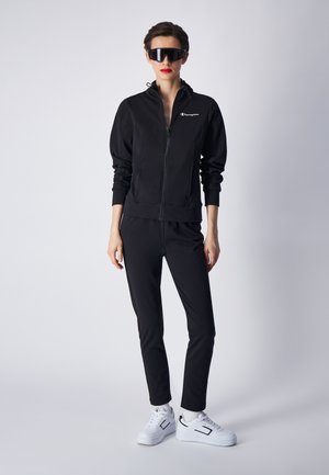 Tuta nera con zip, maniche lunghe e polsini elasticizzati, abbinata a sneakers bianche con accenti neri e suole texturizzate.