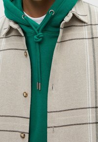 Beige geblokte blouse met grote knopen, gelaagd over een groene hoodie met een trekkoord en een kangoeroezak.