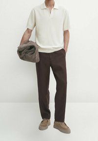 Krèmet kortærmet polo med ribbet struktur, brune tapered bukser og beige sko. Holder en sammenfoldet brun jakke. Minimalistisk baggrund.