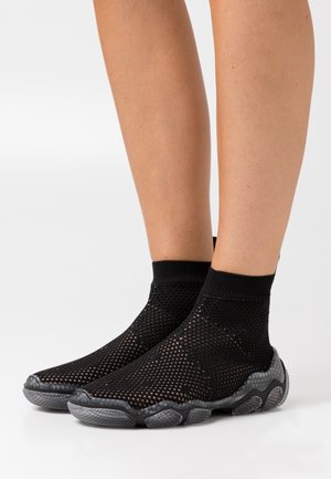 Nohy obuté v černých síťovaných ponožkových botách se strukturovanými šedými podrážkami, stojící na bílém povrchu na jednobarevném pozadí.