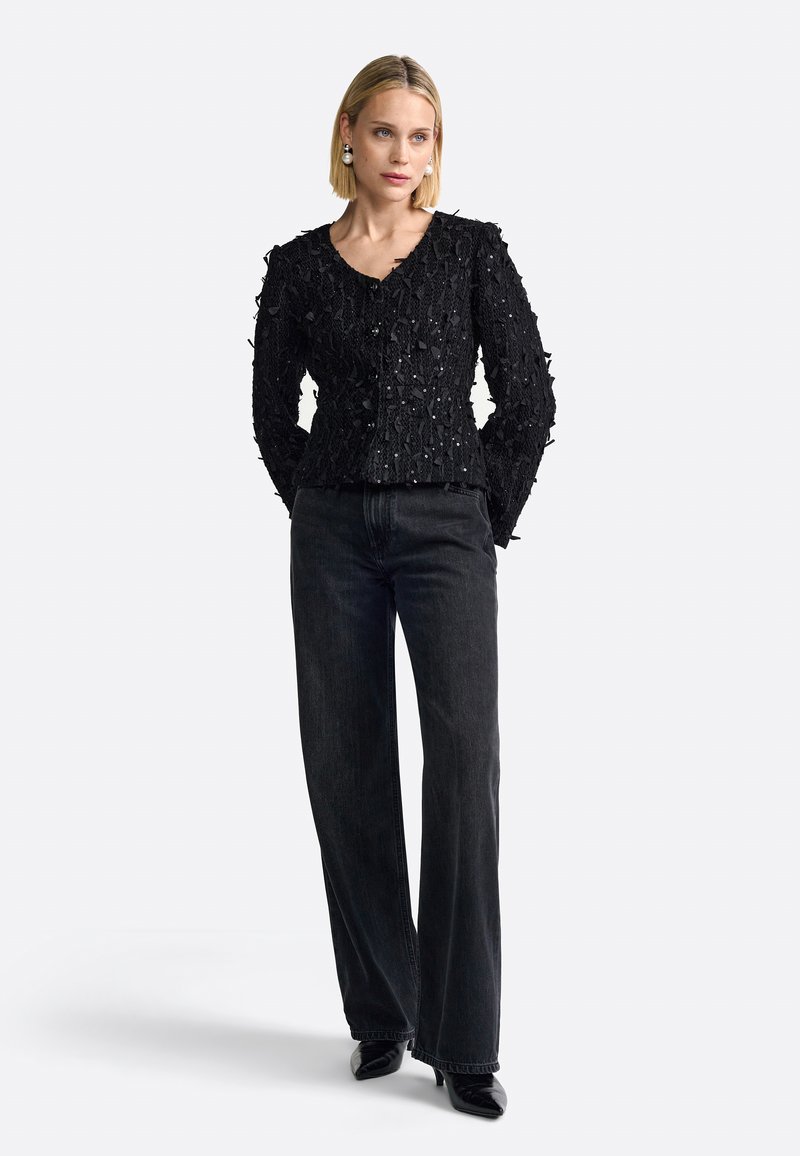 Sorte blondejakke med struktureret mønster og perledekorationer, parret med højtaljede mørke jeans og spidse sorte sko.