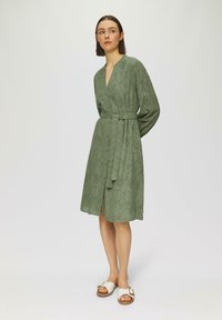 Vestito verde oliva, lungo fino al ginocchio, con scollatura a V, maniche lunghe e vita cinturata. Presenta un motivo ad occhiello e bottoni lungo la parte frontale. Sandali bianchi.