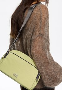 Light green shoulder bag with a smooth texture, zip closure, and gray adjustable strap, worn with a cozy, brown knitted sweater.