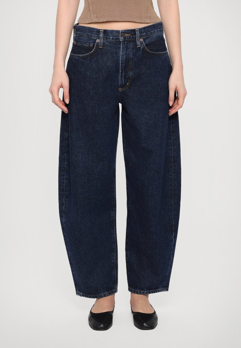 Dunkelblaue Denim-Jeans mit lockerer Passform, hoher Taille, Fünf-Taschen-Design und sichtbaren Nähten. Getragen mit schwarzen Ballerinas.