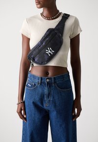 Sac à bandoulière à motif camouflage en bleu foncé, avec un logo blanc. Sangle réglable, fermeture éclair, fabriqué en matériau durable, porté en travers du corps.