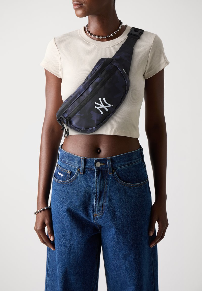 Sac à bandoulière à motif camouflage en bleu foncé, avec un logo blanc. Sangle réglable, fermeture éclair, fabriqué en matériau durable, porté en travers du corps.