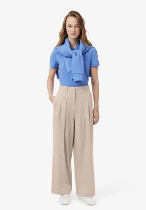 Donna che indossa una polo azzurra con maglione abbinato sulle spalle, pantaloni beige a gamba larga e sneaker bianche, in piedi.