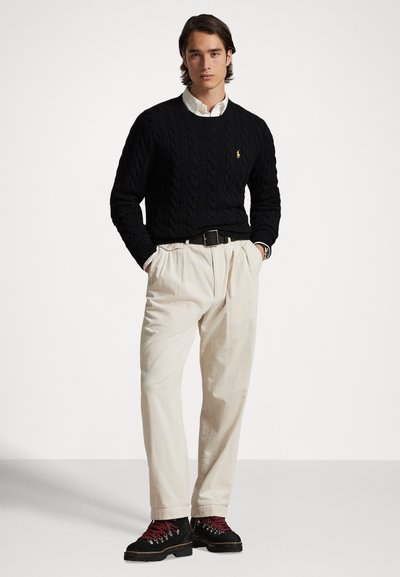 Άνδρας που φοράει μαύρο πουλόβερ με πλεκτό σχέδιο καλωδίων, λευκό πουκάμισο με γιακά, μπεζ πτυχωτό παντελόνι, μαύρη ζώνη και μαύρες μπότες με κόκκινες κορδόνια, στέκεται με τα χέρια στις τσέπες.