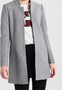 Grå, skräddarsydd kappa med hög krage och långa ärmar. Tillverkad av ett strukturerat tyg, med sidofickor och en rak siluett.